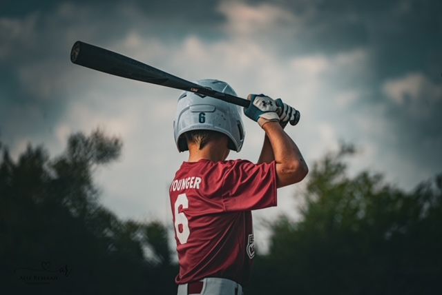 Baseball player in red jersey and white helmet takes batting stance against moody cloudy sky.