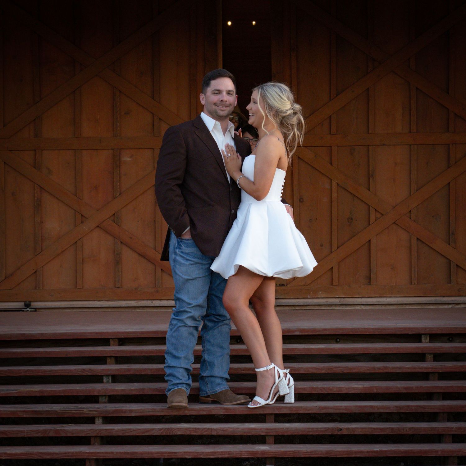 Two pairs of feet in dress shoes stand on wooden steps against a rustic barn wood background at dusk.