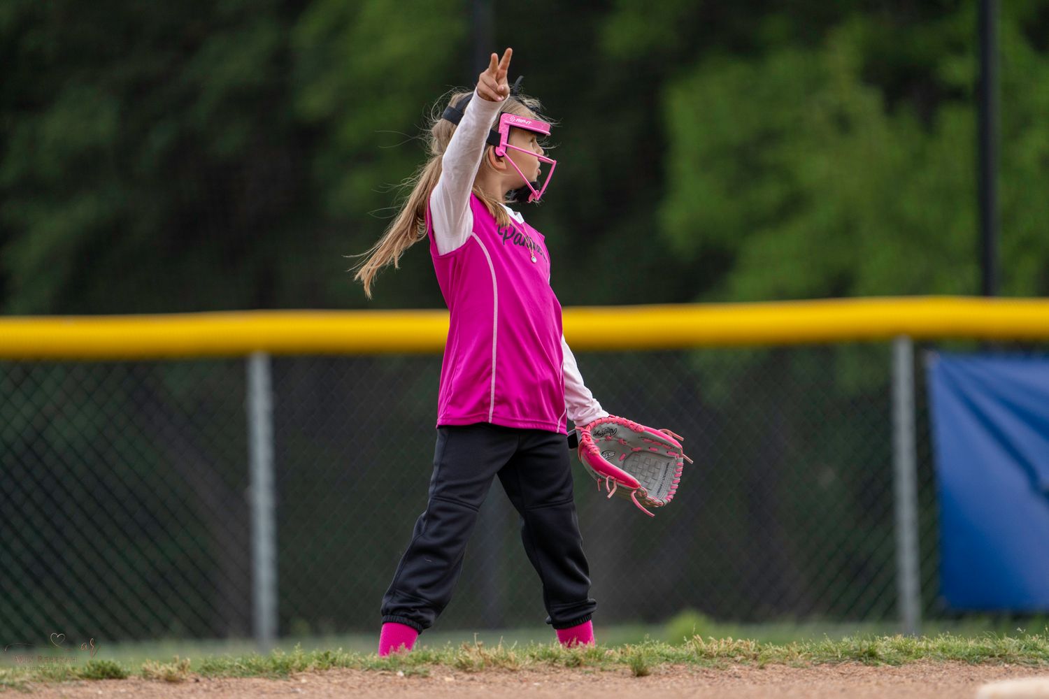 Young softball pitcher in pink uniform winds up to throw a pitch during a game on a sunny day.