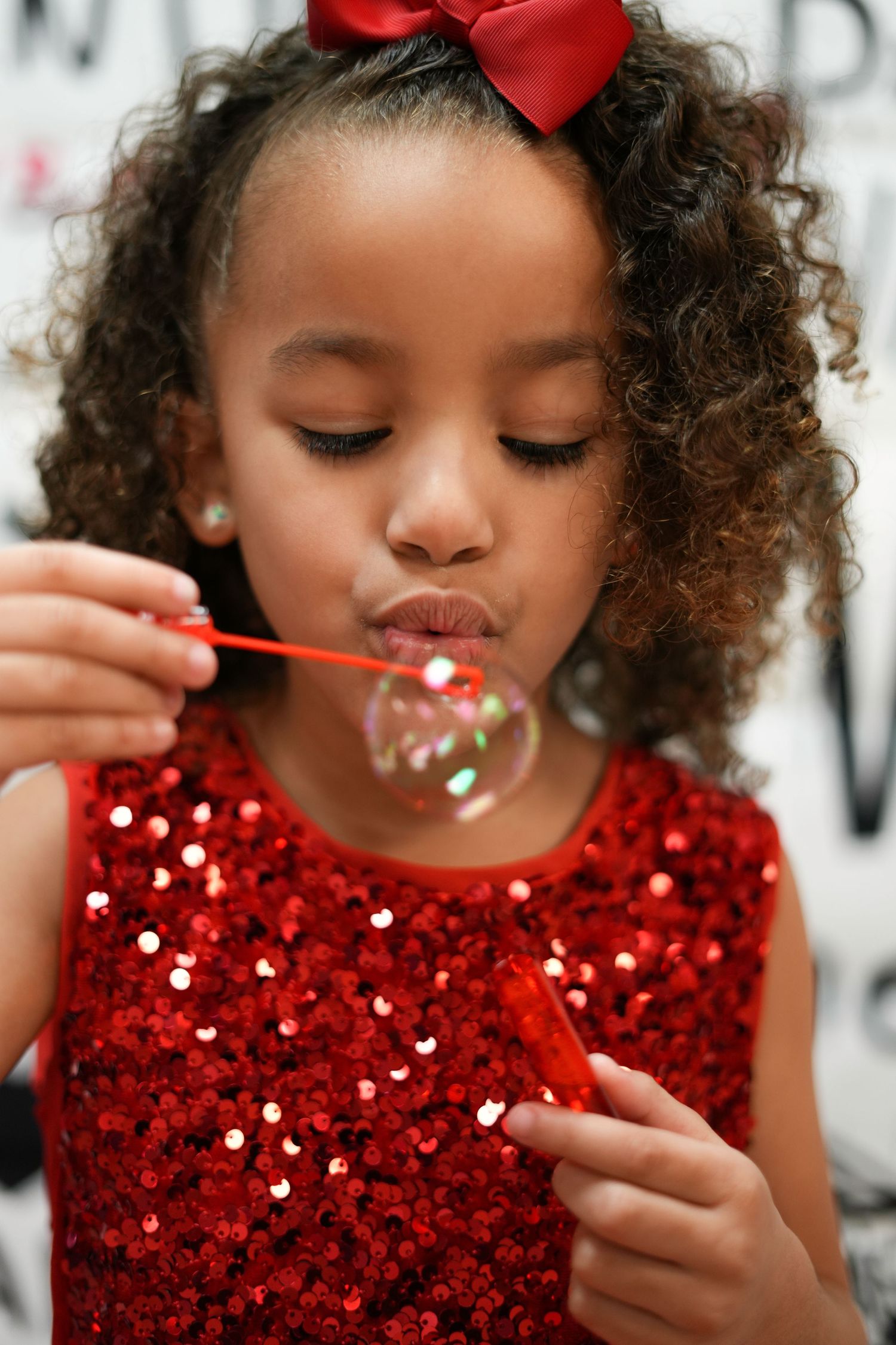 Child in red sequined dress blowing bubbles with eyes closed and red bow in curly hair.