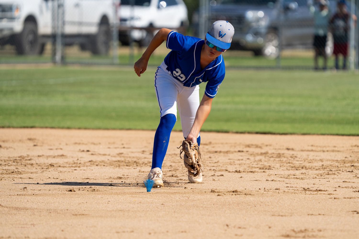 Baseball player in blue uniform bends down to field a ground ball during game on dirt infield.
