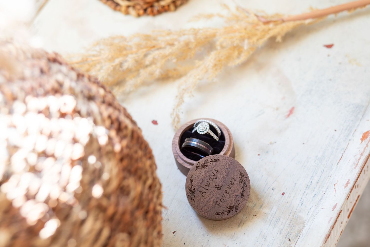 A diamond engagement ring displayed in an open wooden ring box with dried pampas grass on a rustic white surface.