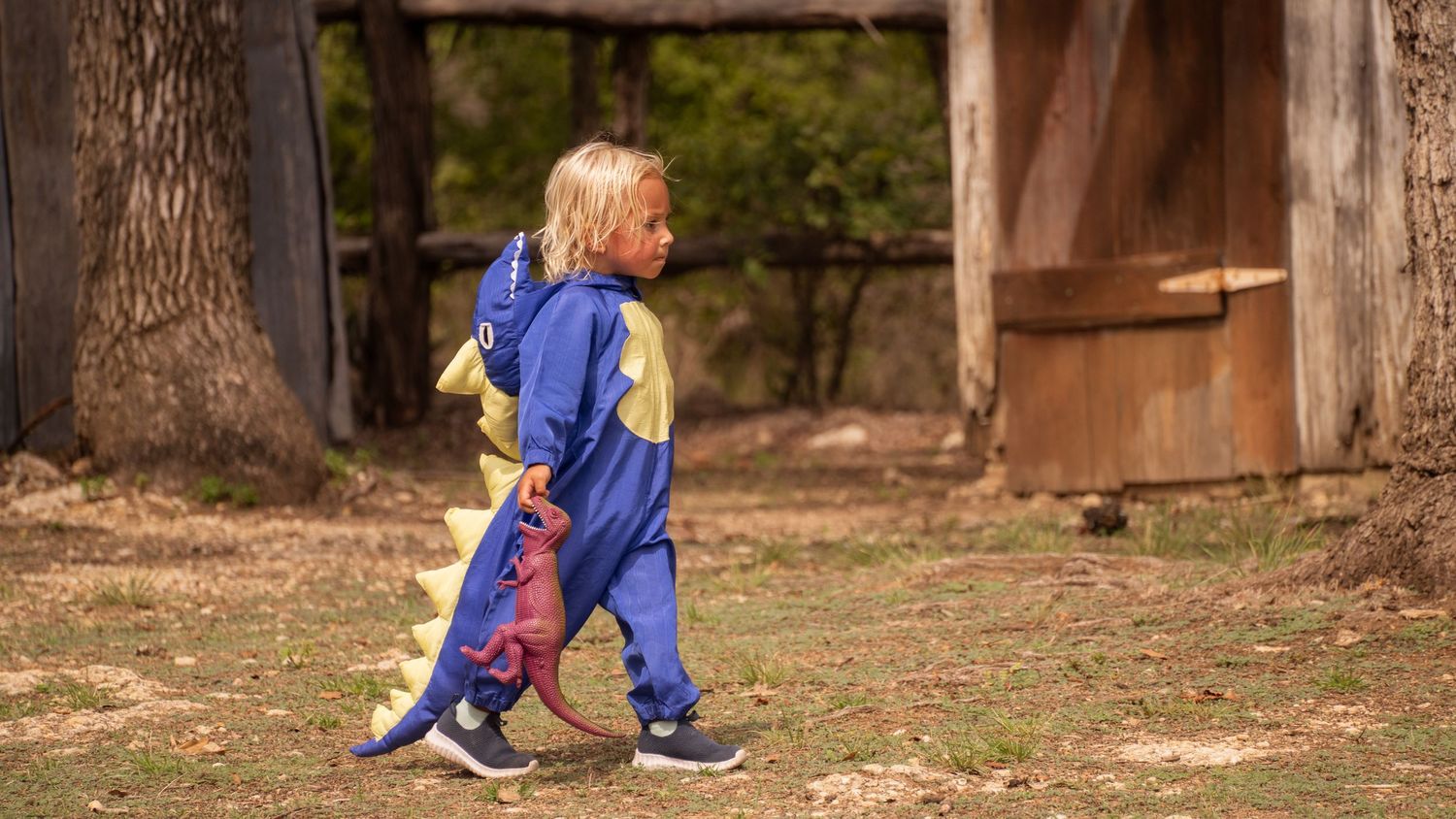 A toddler walking outdoors wearing blue overalls in a rustic setting.