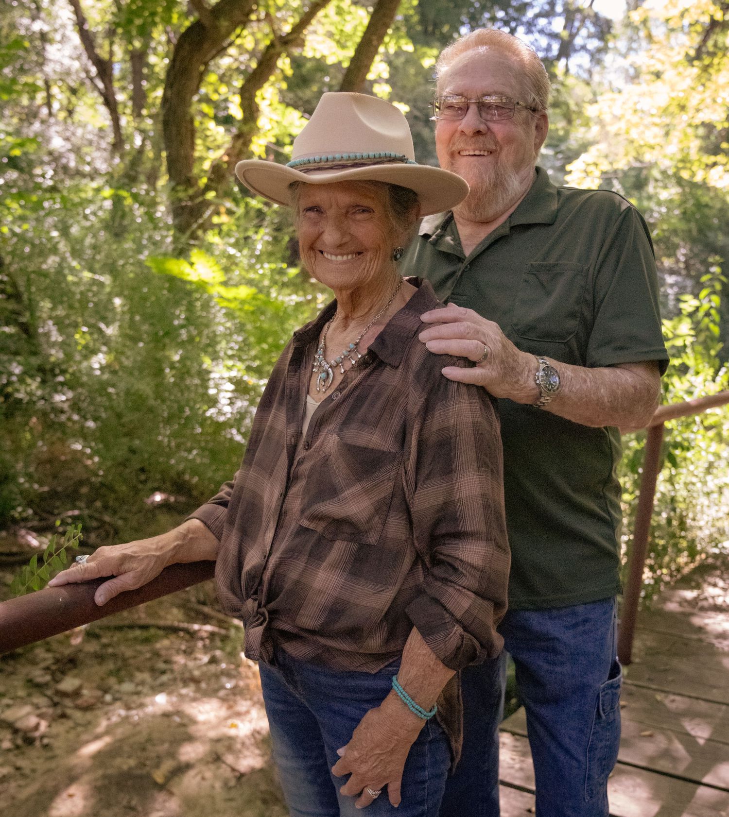 A couple wearing casual western attire shares a tender moment outdoors in a sun-dappled forest setting.