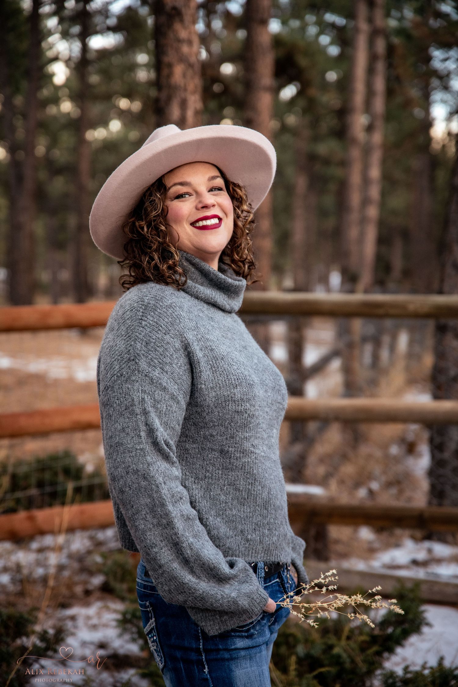 Winter outdoor portrait of woman in grey sweater and light hat against snowy forest backdrop.