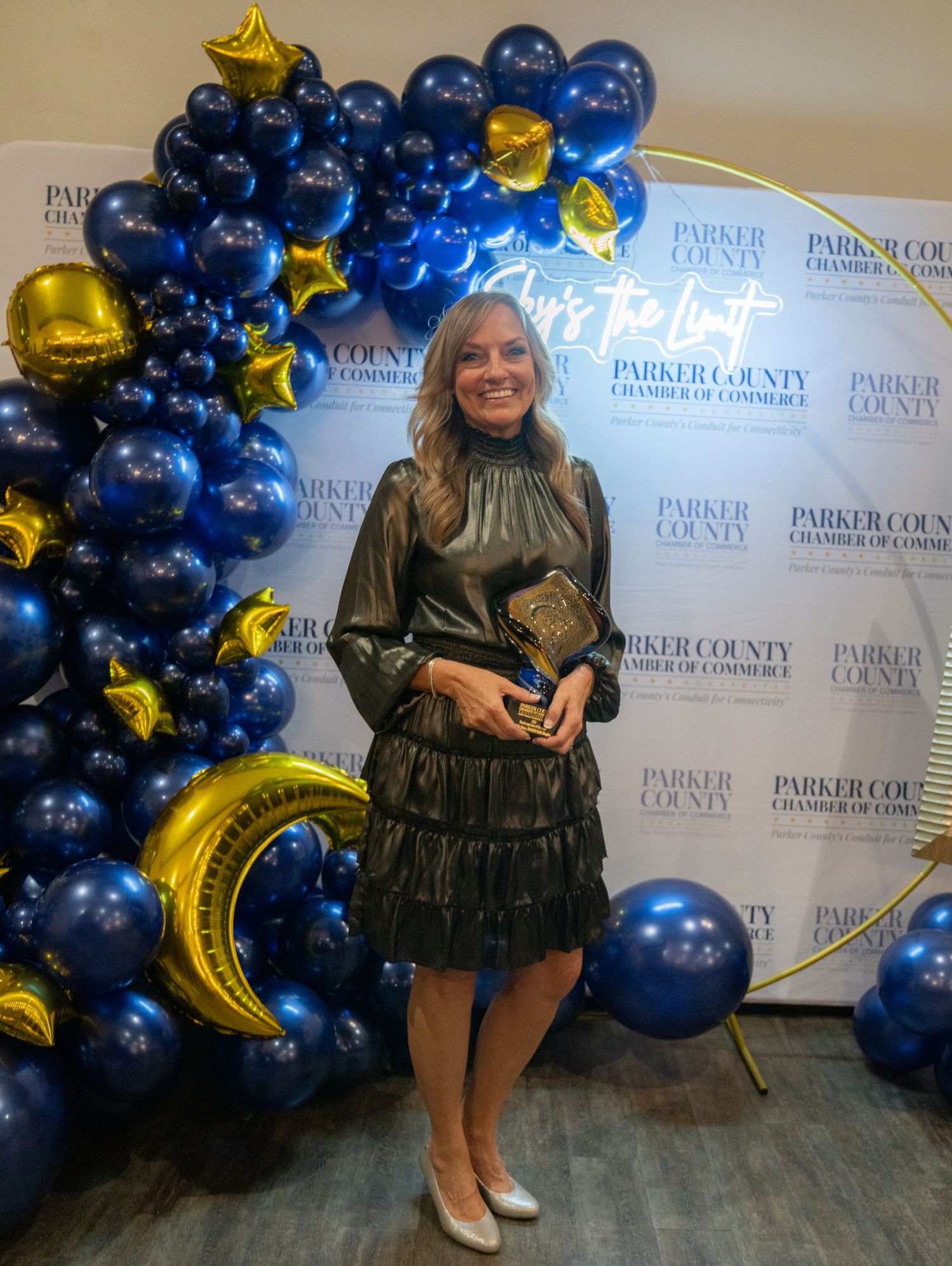 Award winner poses with trophy against blue balloon arch with gold stars at Parker County Chamber event.