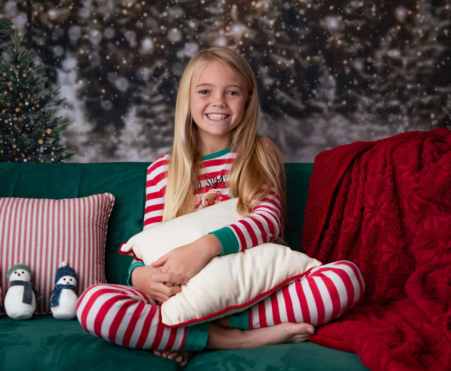 A child in striped Christmas pajamas sits on a green couch with holiday pillows.