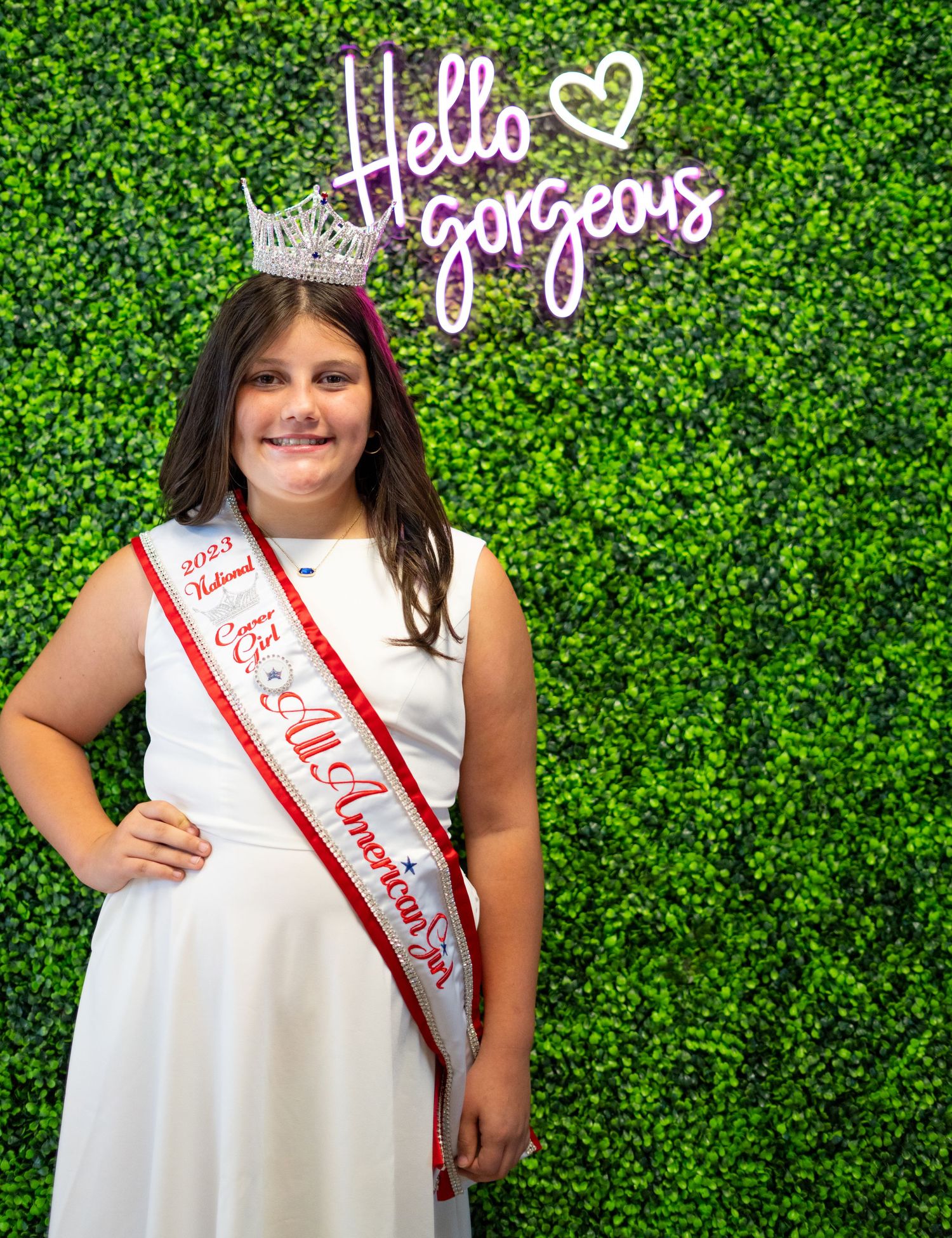 Pageant winner in white dress poses against green hedge wall with 'Hello Gorgeous' text and sparkly crown.