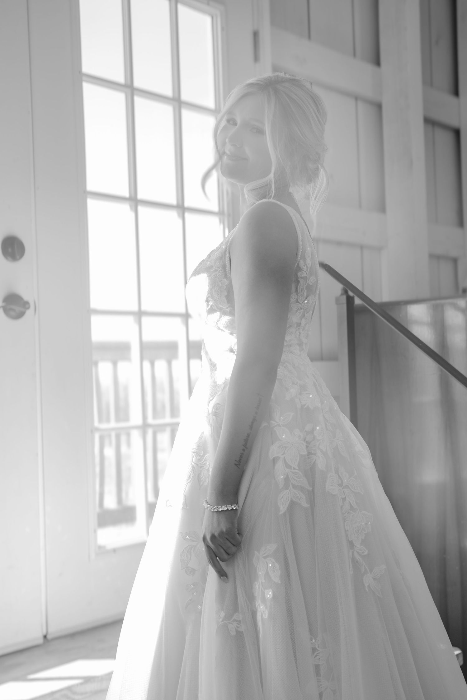 A backlit silhouette shows a bride in a flowing lace wedding gown standing by a bright window in black and white.