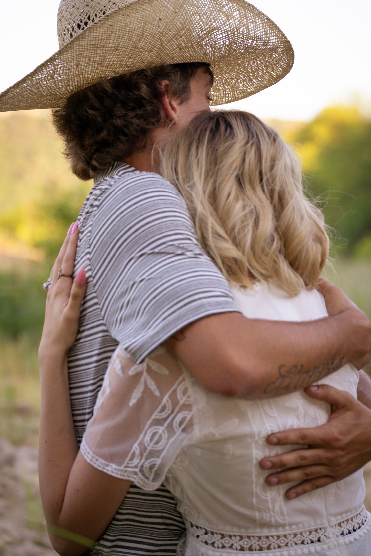 A couple embraces outdoors at sunset, with one person wearing a straw hat and striped shirt.