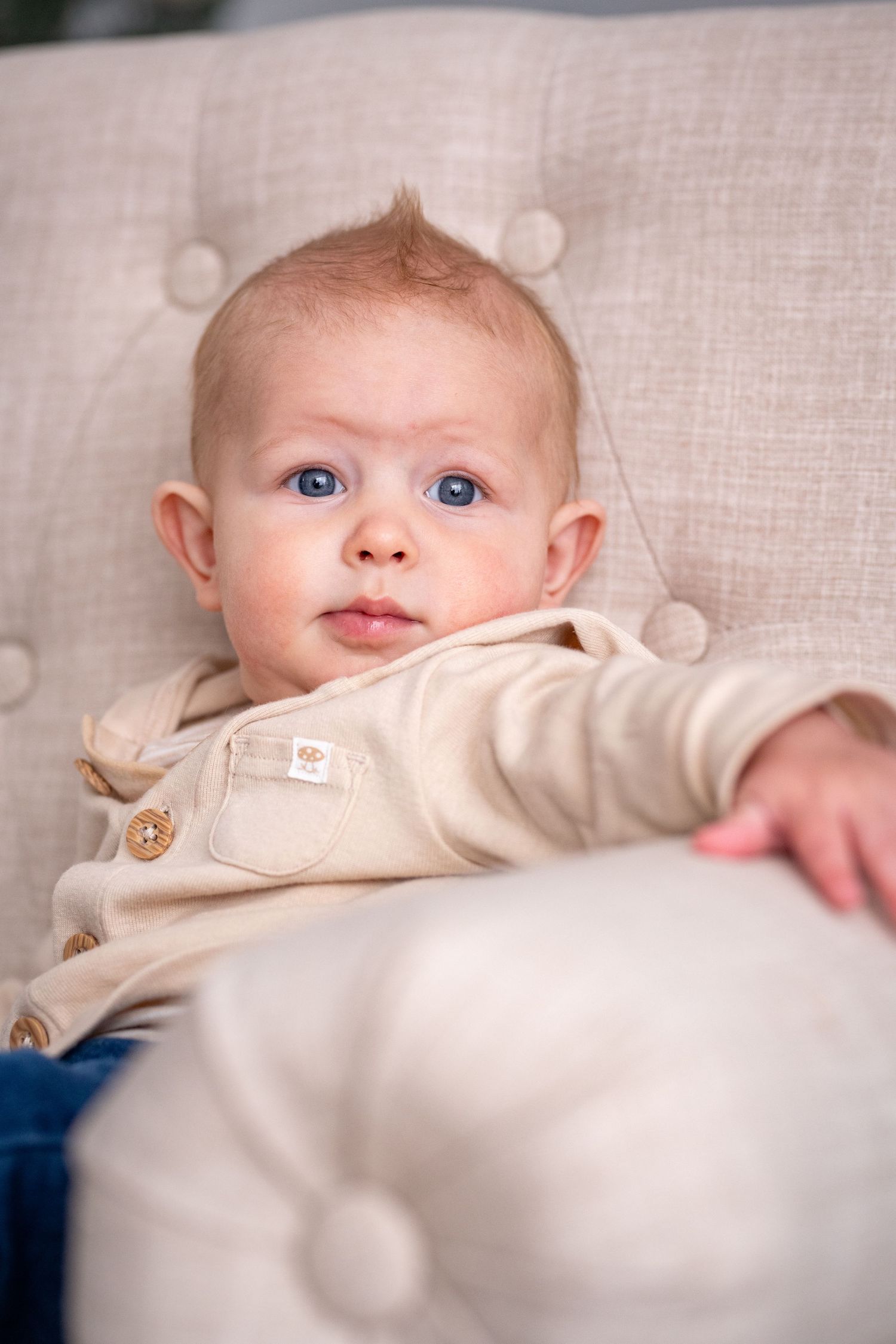 A young baby in a beige sweater with buttons rests on light-colored furniture while gazing upward with bright blue eyes.