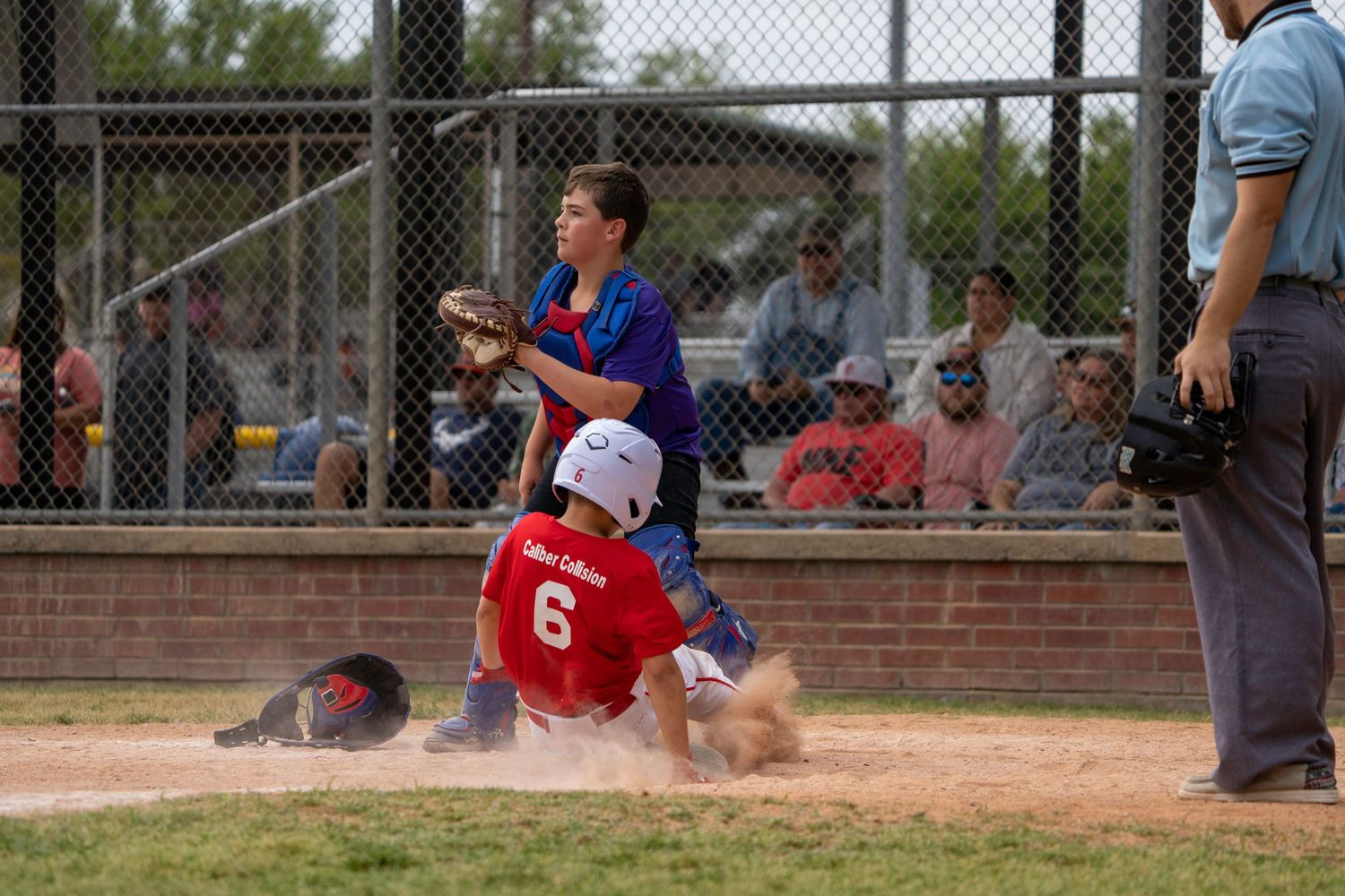 Youth baseball player slides into home plate as catcher in blue uniform tries to make the tag during game.