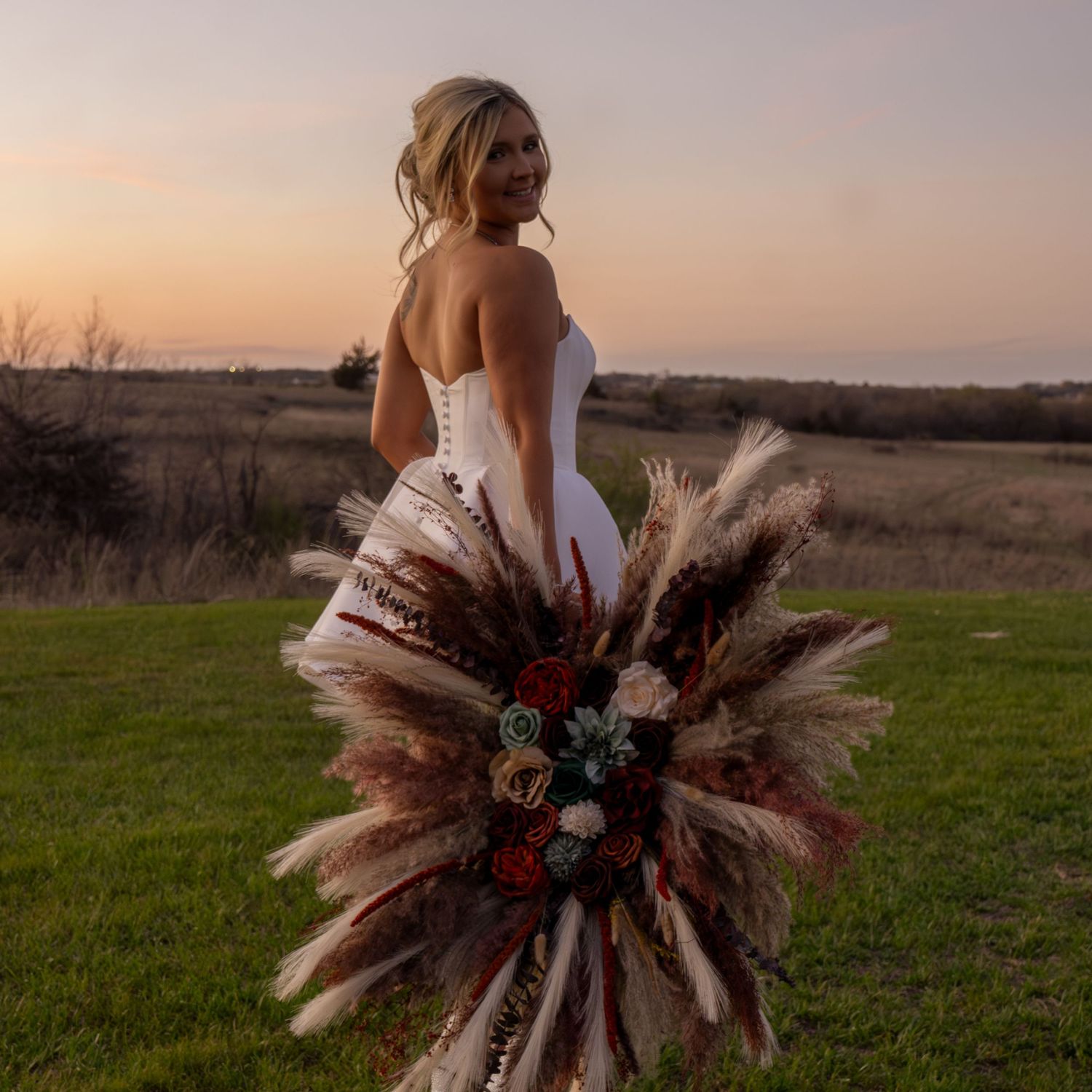 Person in white wedding dress holding large dried pampas grass and floral bouquet at sunset in grassy field.