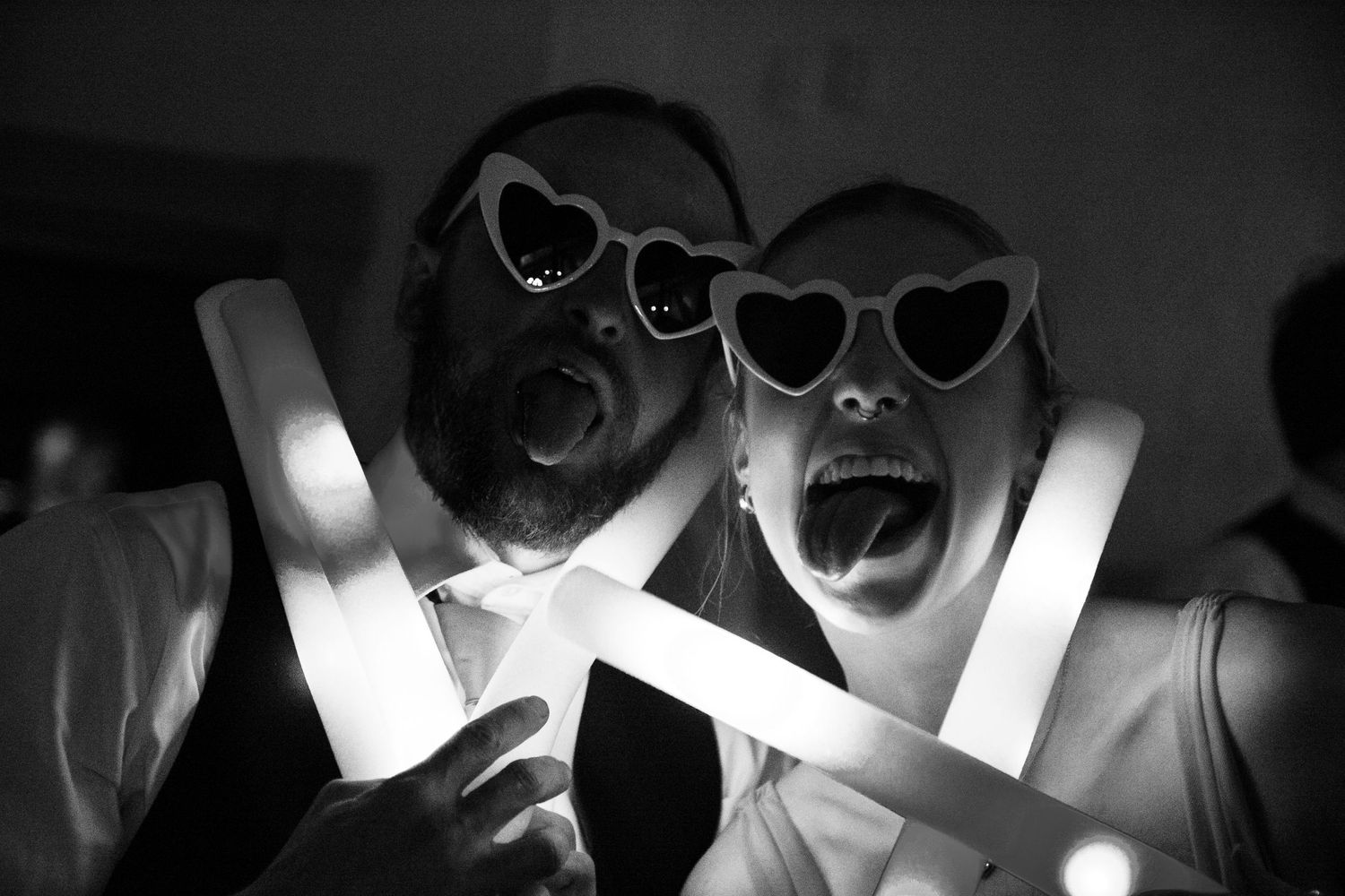 Two party-goers wearing heart-shaped sunglasses hold glowing light sticks in black and white.