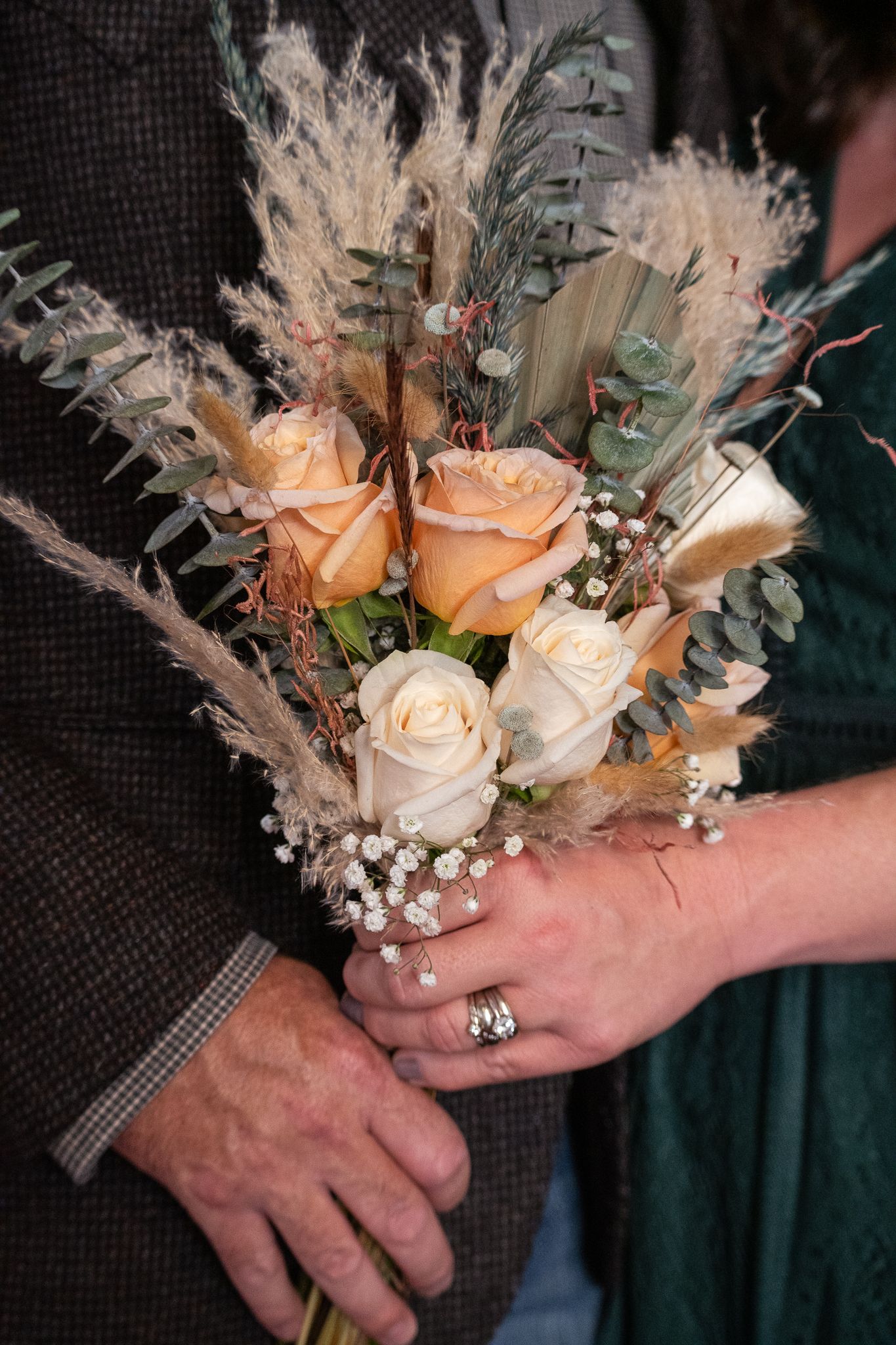 Close-up of hands holding peach and cream rose bouquet with dried flowers.