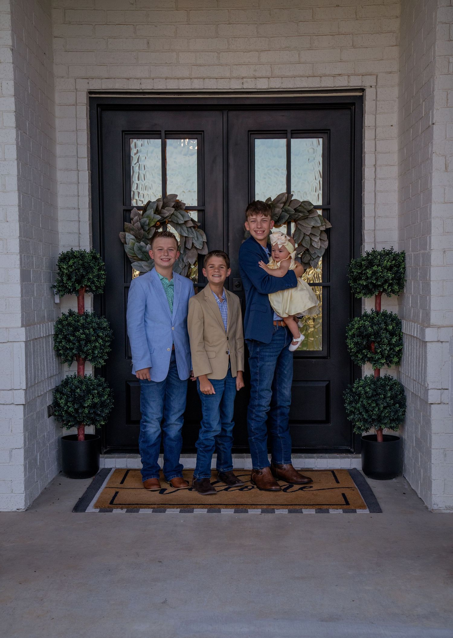 A group stands on a front porch with a black double door, flanked by decorative topiary plants and holiday wreaths.