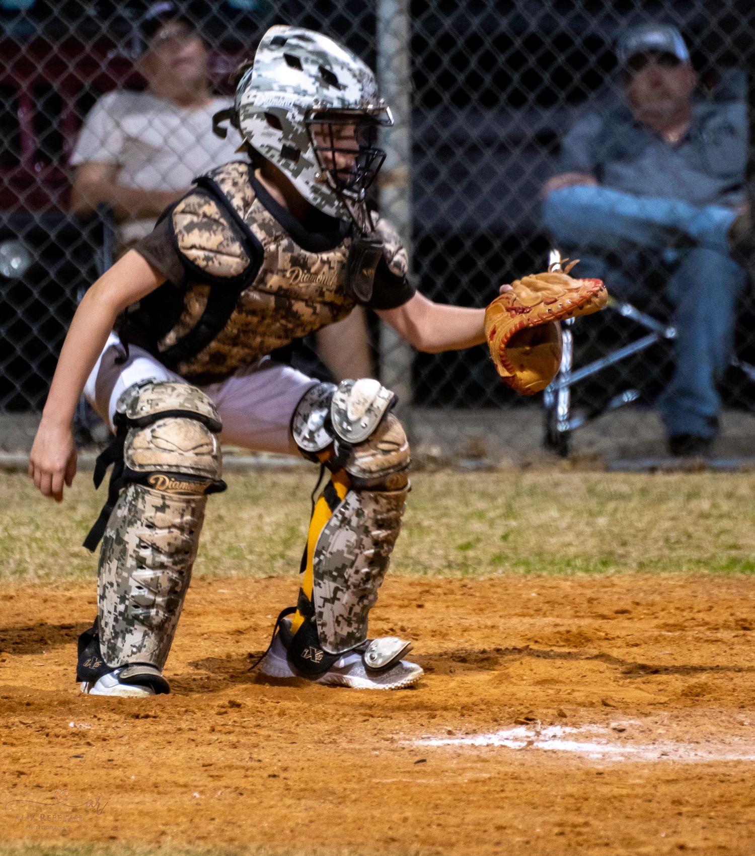 Baseball catcher in camouflage gear crouches in ready position at home plate during a game.
