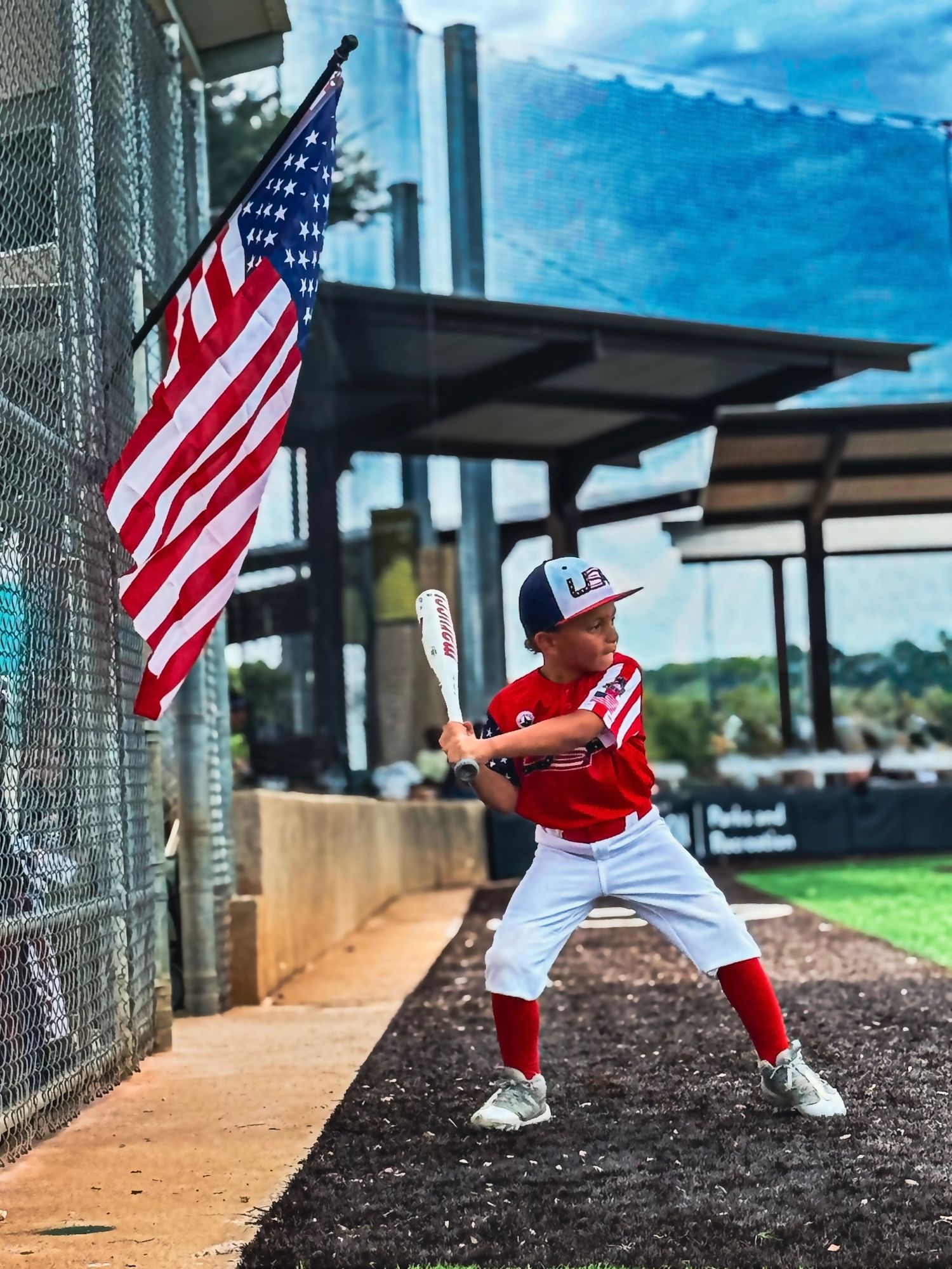 Young baseball player in USA uniform holds bat in batting stance next to American flag at outdoor baseball field.