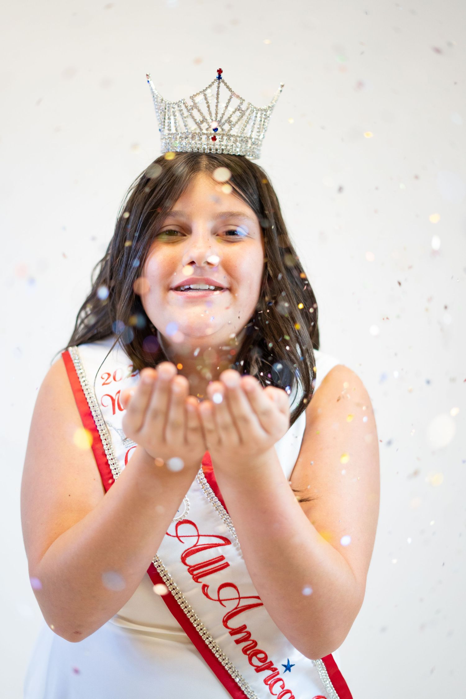 Pageant winner in white dress blowing sparkly confetti while wearing crown and sash.