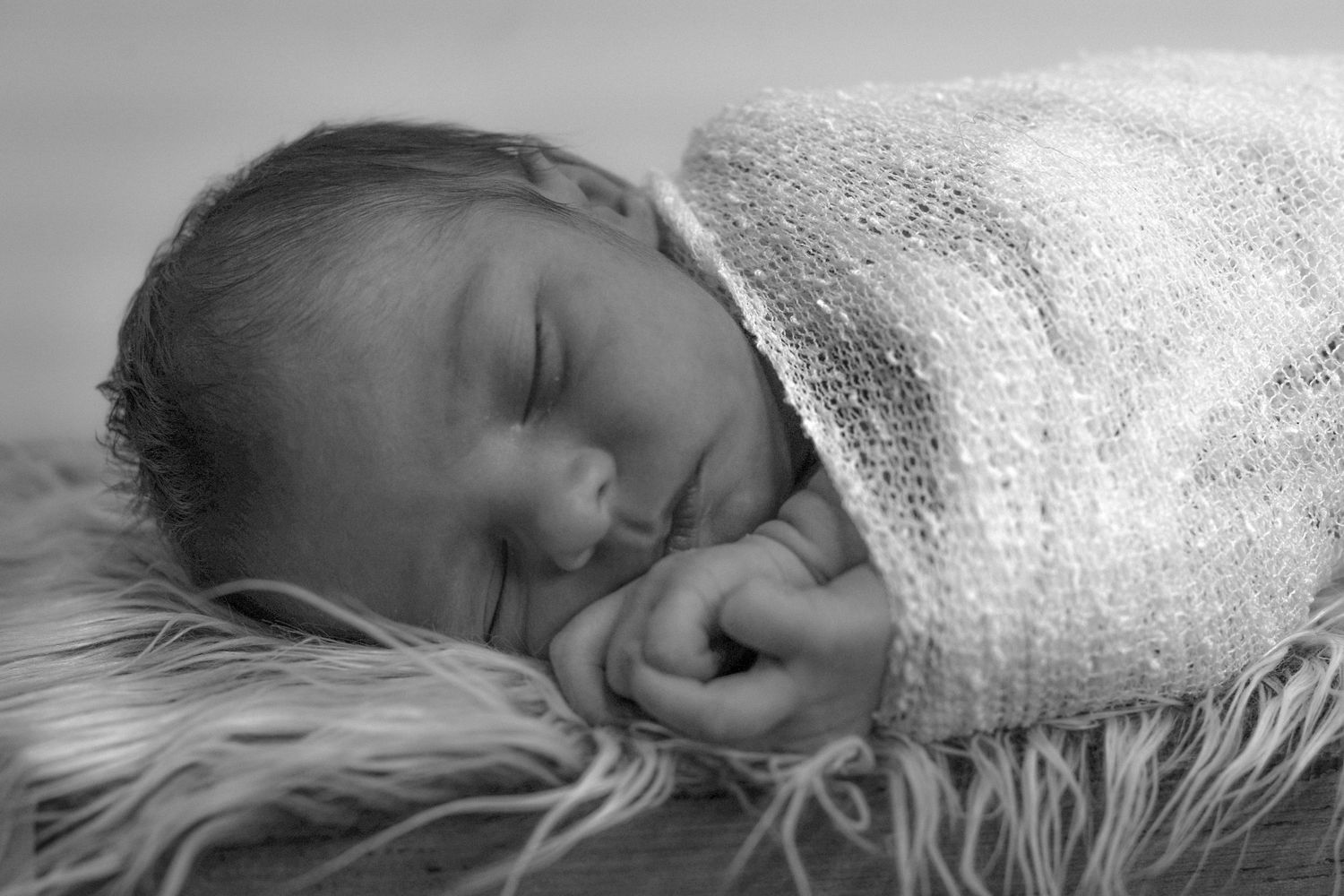 Sleeping newborn wrapped in soft white blanket on furry backdrop in black and white.