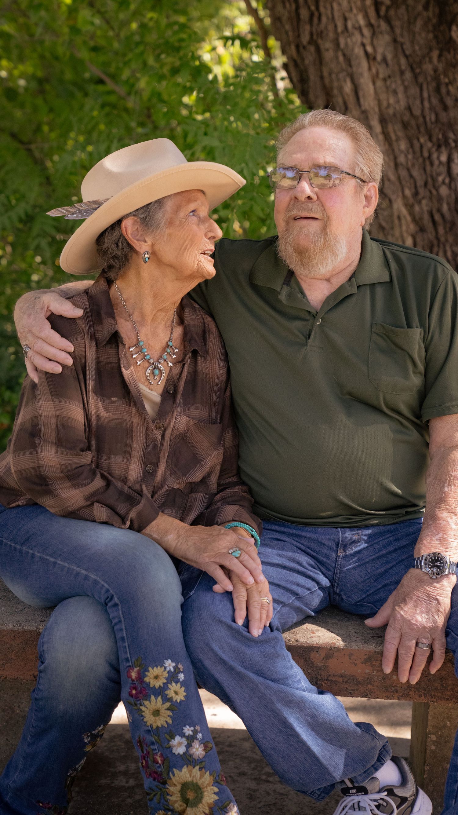 A couple sits together on a wooden bench surrounded by trees in a peaceful outdoor setting.