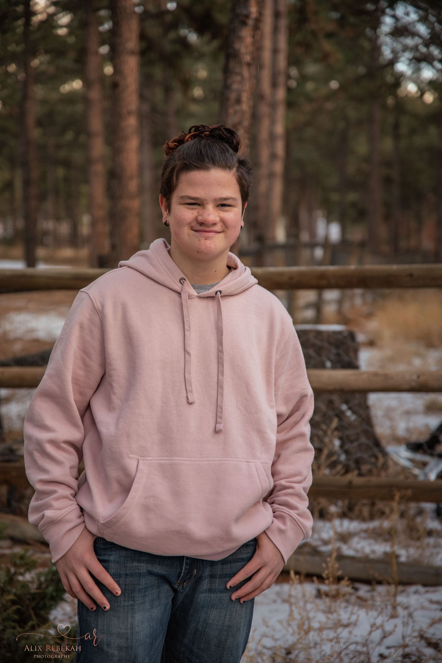 Person in pink hoodie smiling outdoors with pine trees and wooden fence in background.