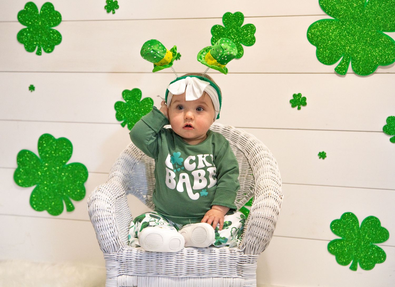 Baby in St Patrick's Day outfit sitting in white chair surrounded by shamrock decorations.