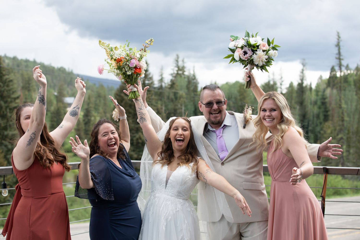 Wedding party celebrates joyfully on outdoor deck with mountain scenery.