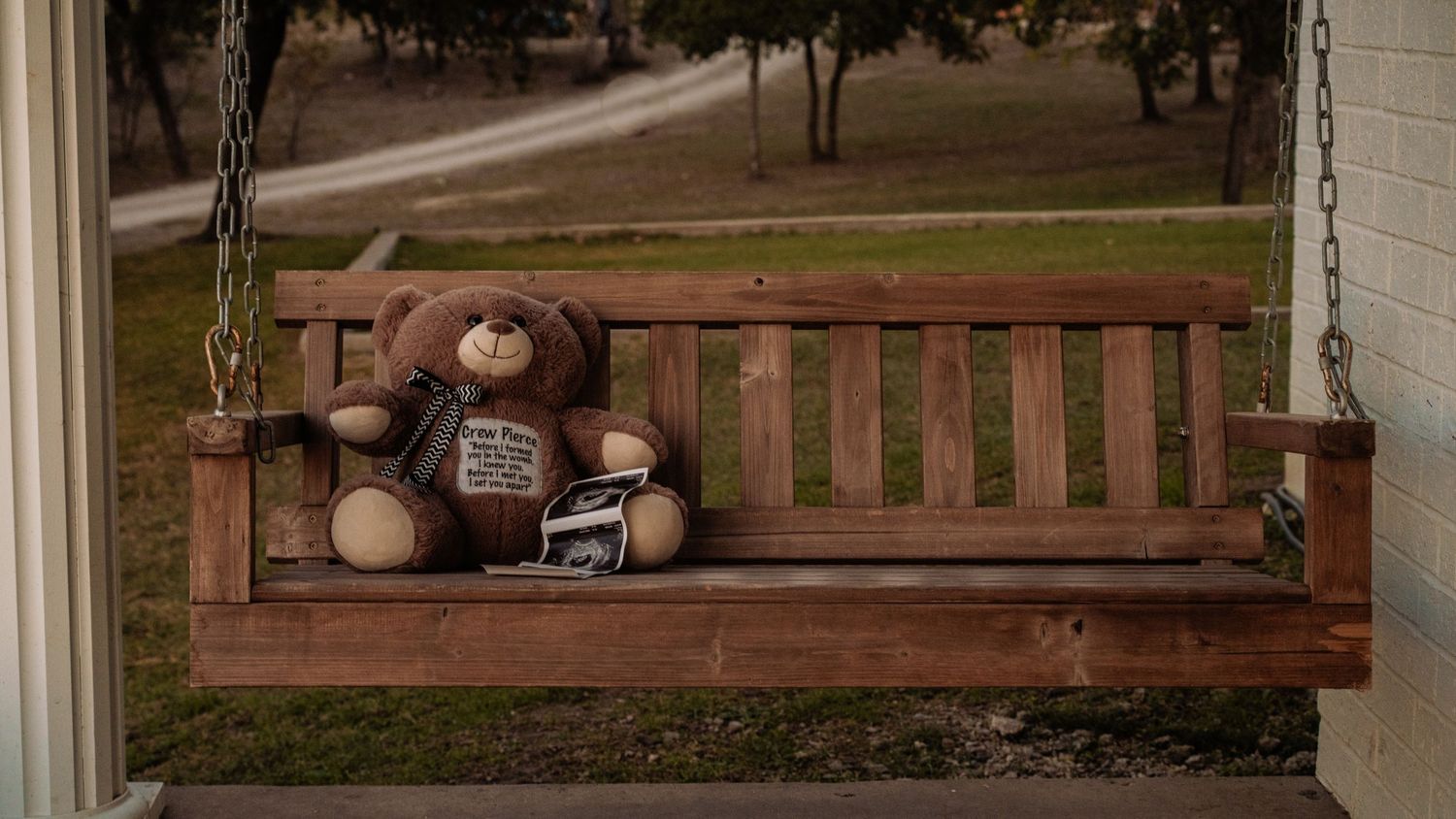 Teddy bear with ultrasound photo sits alone on wooden porch swing.