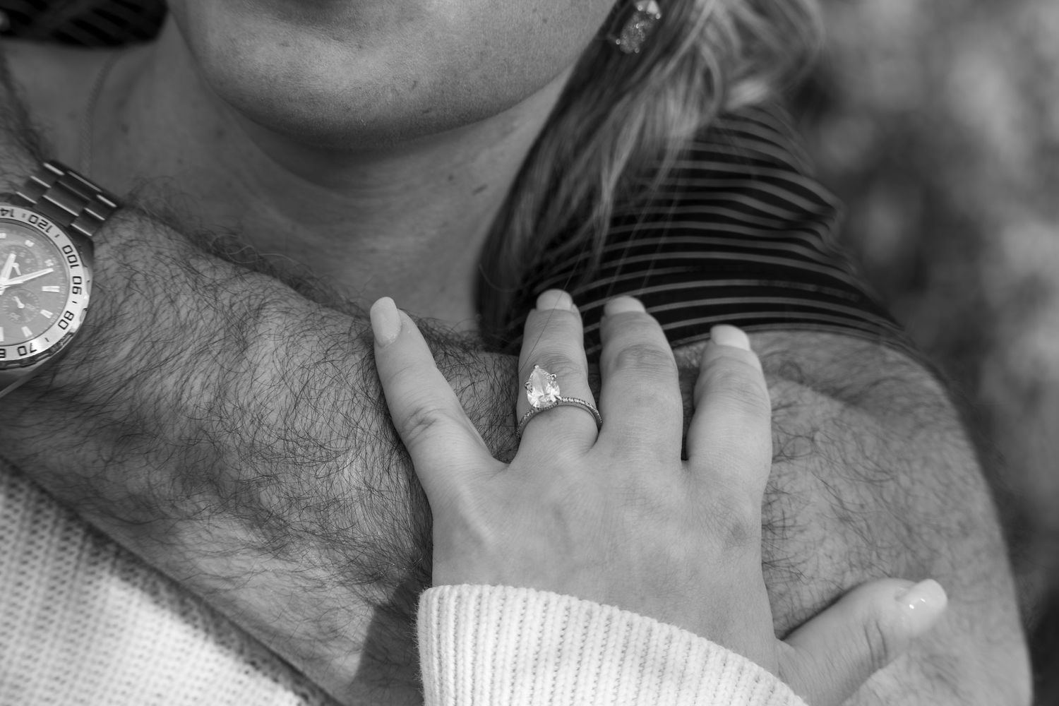 Black and white photo of a couple embracing, showcasing an engagement ring and a watch on their wrists.