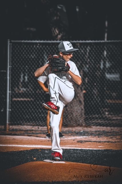A baseball pitcher in white uniform and cap winds up for a pitch on the pitching mound against a chain-link fence background.
