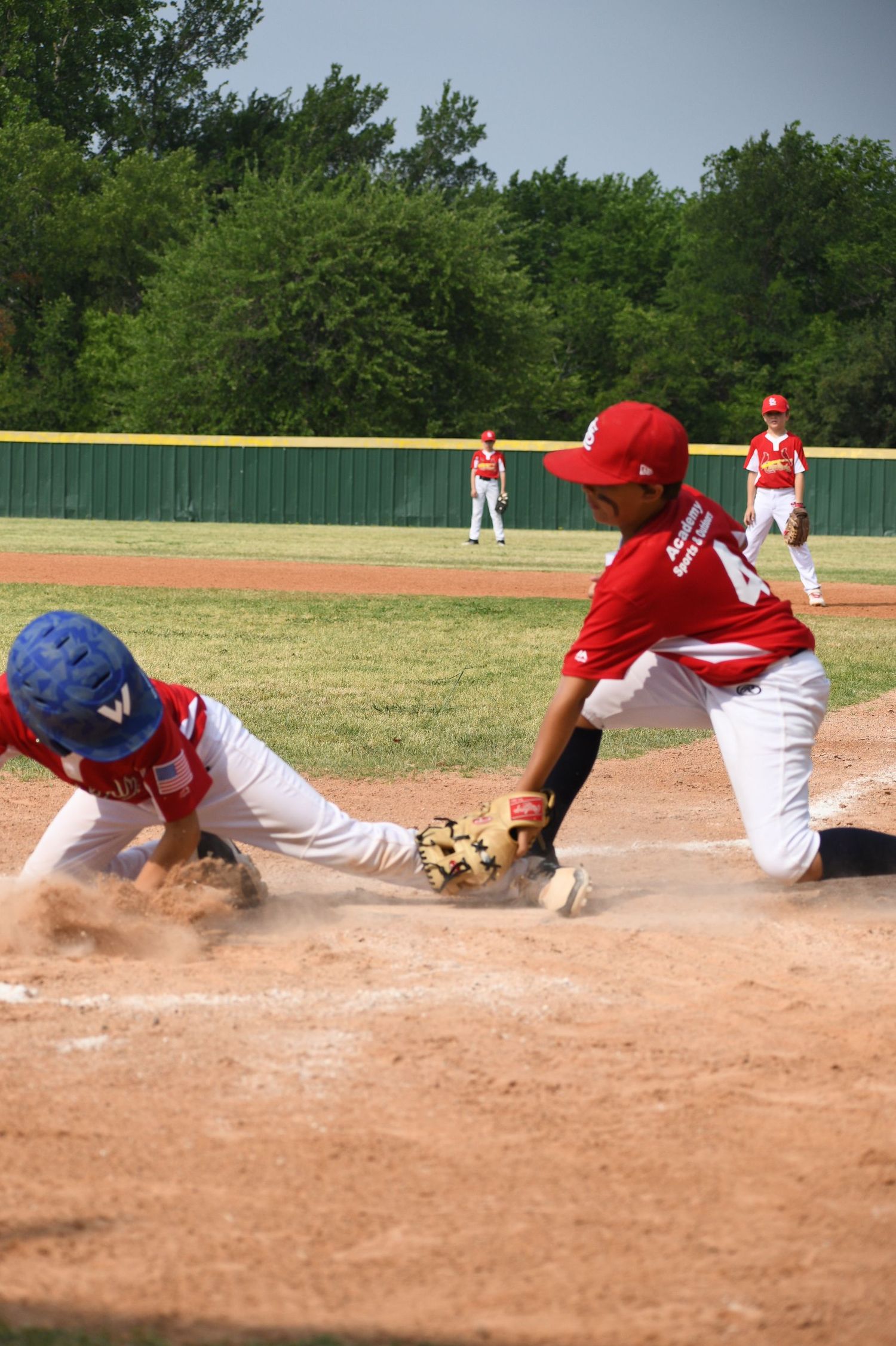 Baseball players in red and blue uniforms compete for the ball during a close play at home plate on a dirt field.