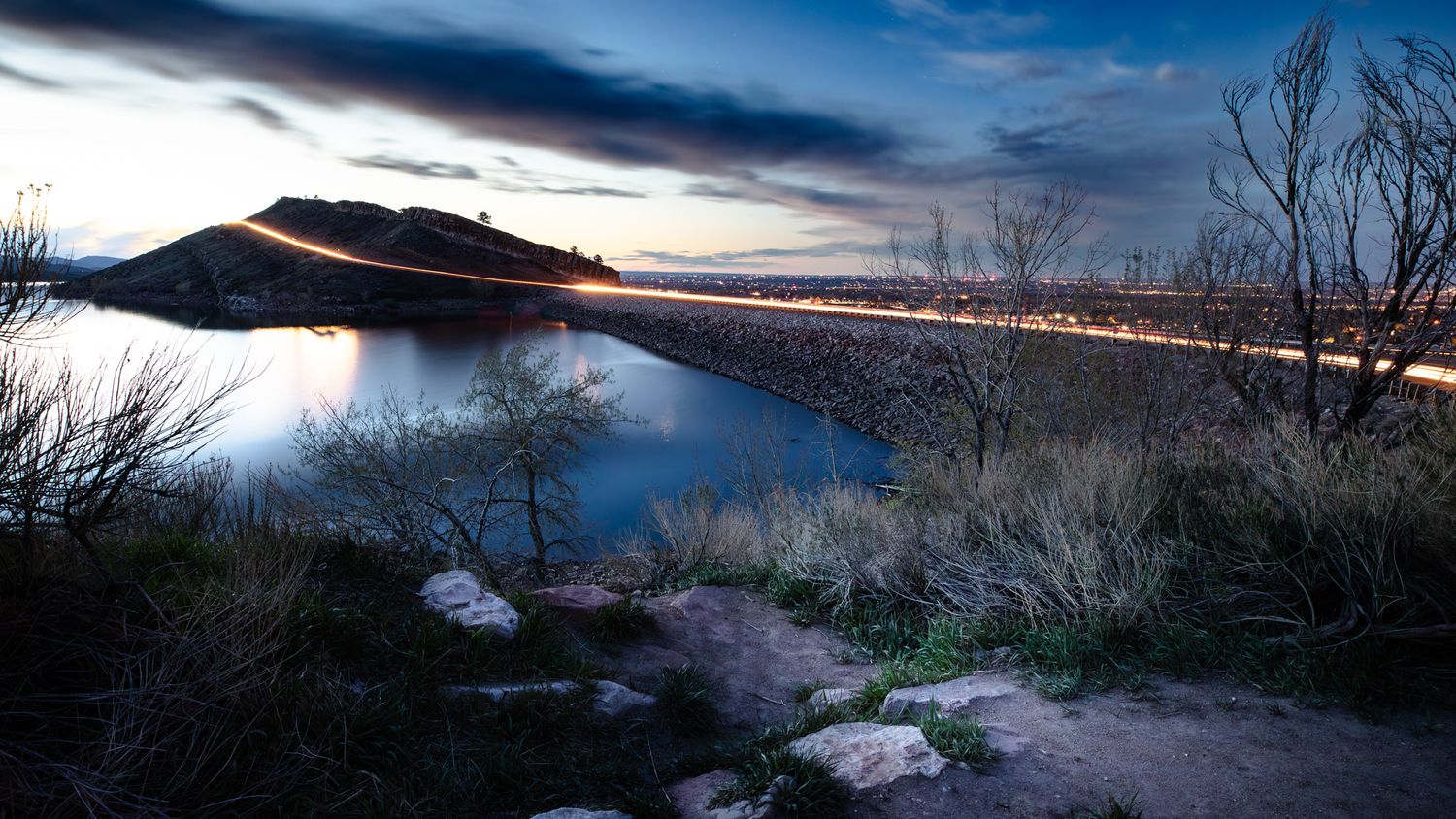 Long exposure of car lights streaking along lakeside road at twilight with dramatic cloudy sky.