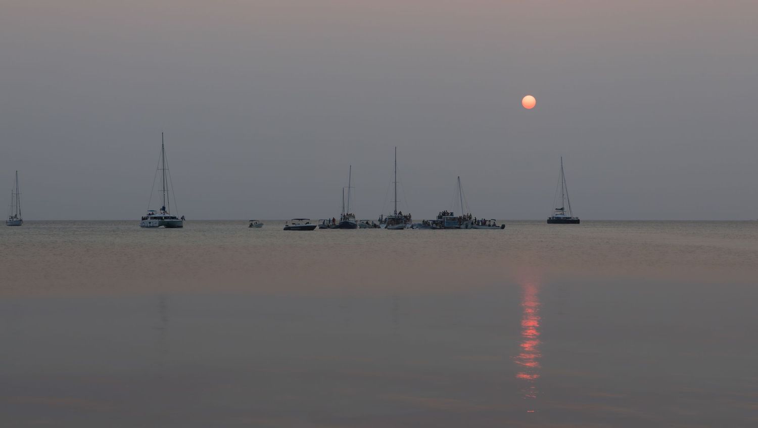 Sailboats silhouetted against a hazy sunset over calm waters with orange sun reflecting on the surface.