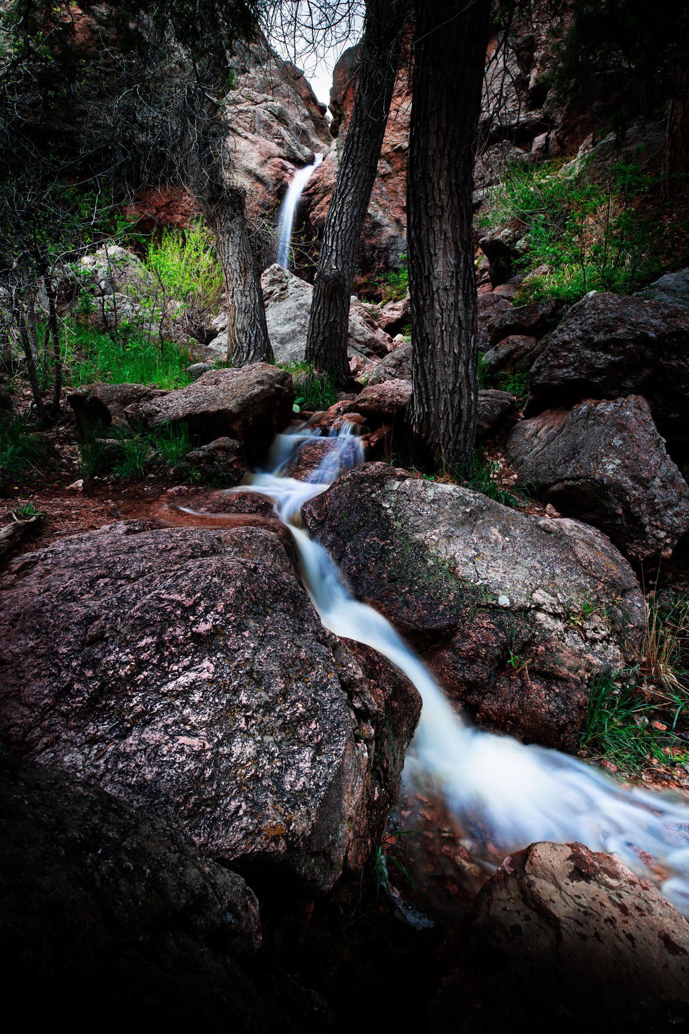 Cascading waterfall flowing over rocky terrain between dark tree trunks in a forest setting.