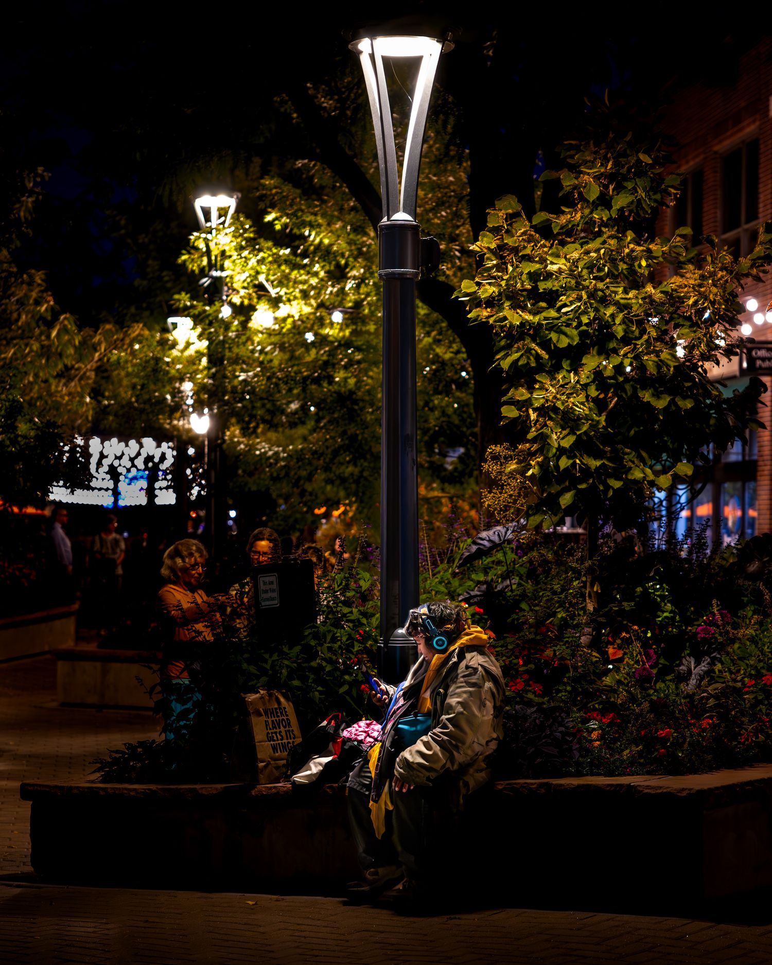 Person sitting under illuminated street lamp at night surrounded by trees and urban scenery.