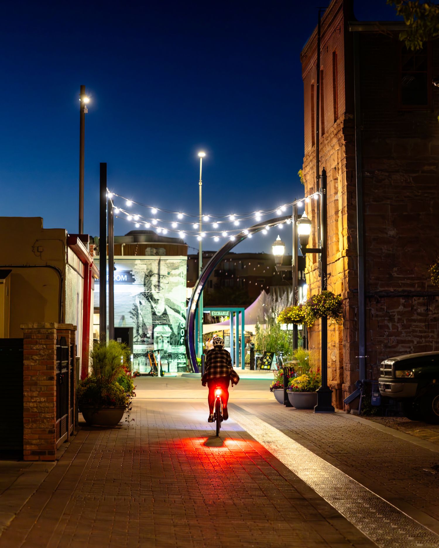 Cyclist on illuminated street at night with string lights and storefronts creating ambient glow.