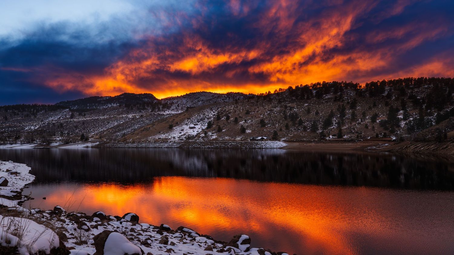 Dramatic sunset over mountain lake with vibrant orange clouds reflecting on water.