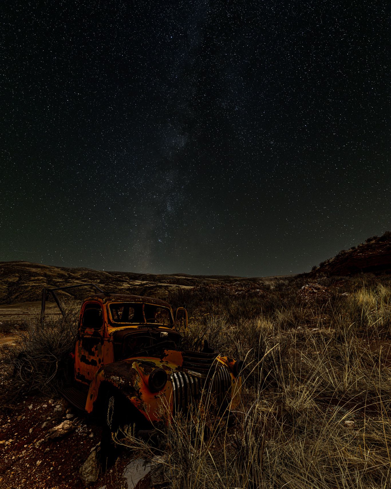 Starry night sky over a rustic abandoned car in a desert field.
