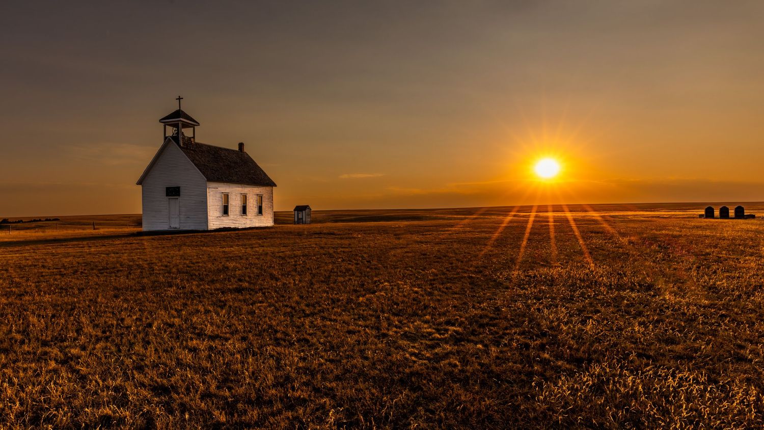 Small white church stands silhouetted against a golden prairie sunset.