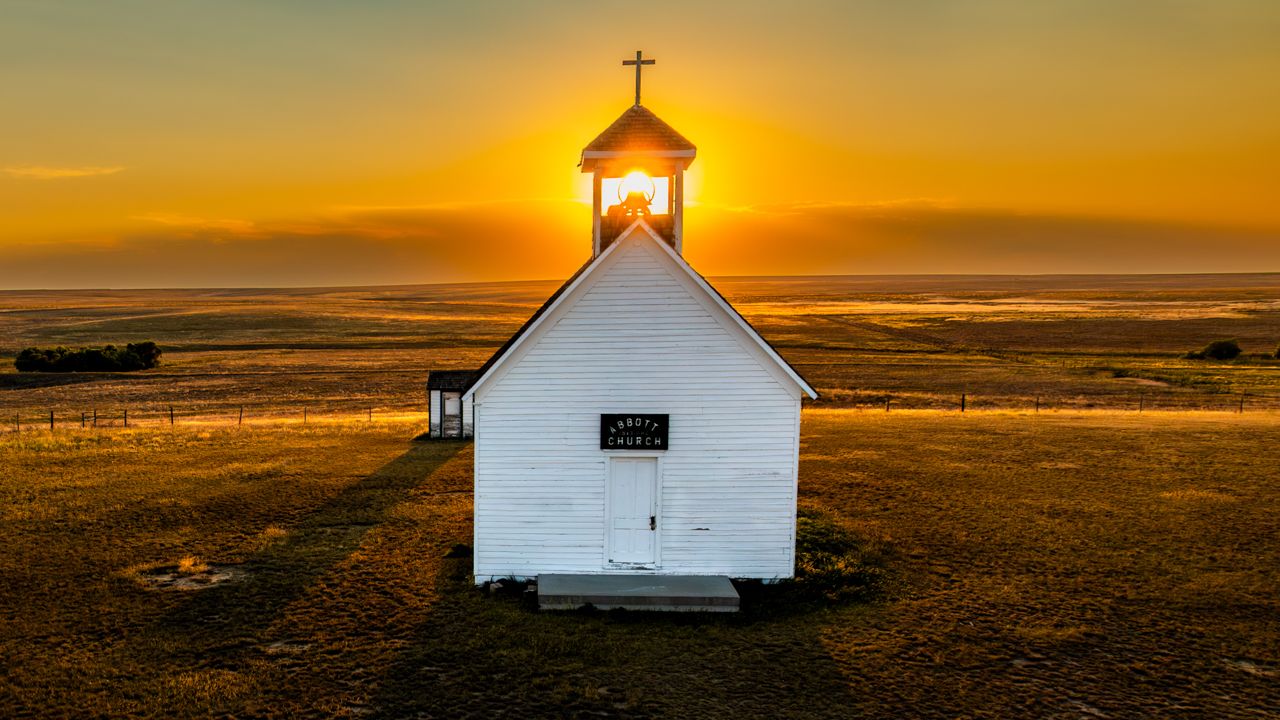 White country church backlit by golden sunset shining through steeple window.