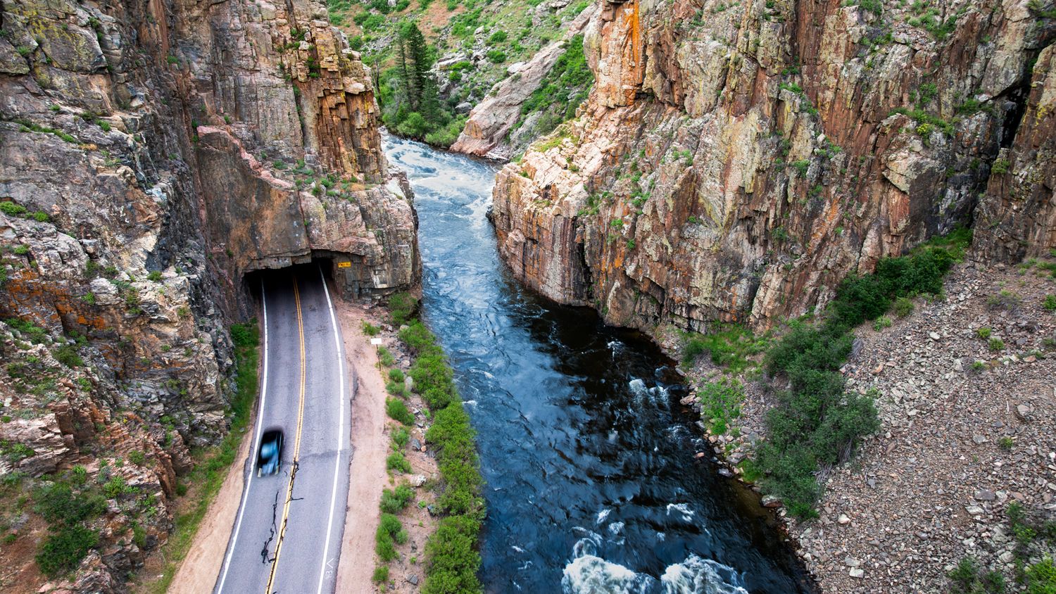 Winding mountain road follows a river through a dramatic rocky canyon with steep cliffs.