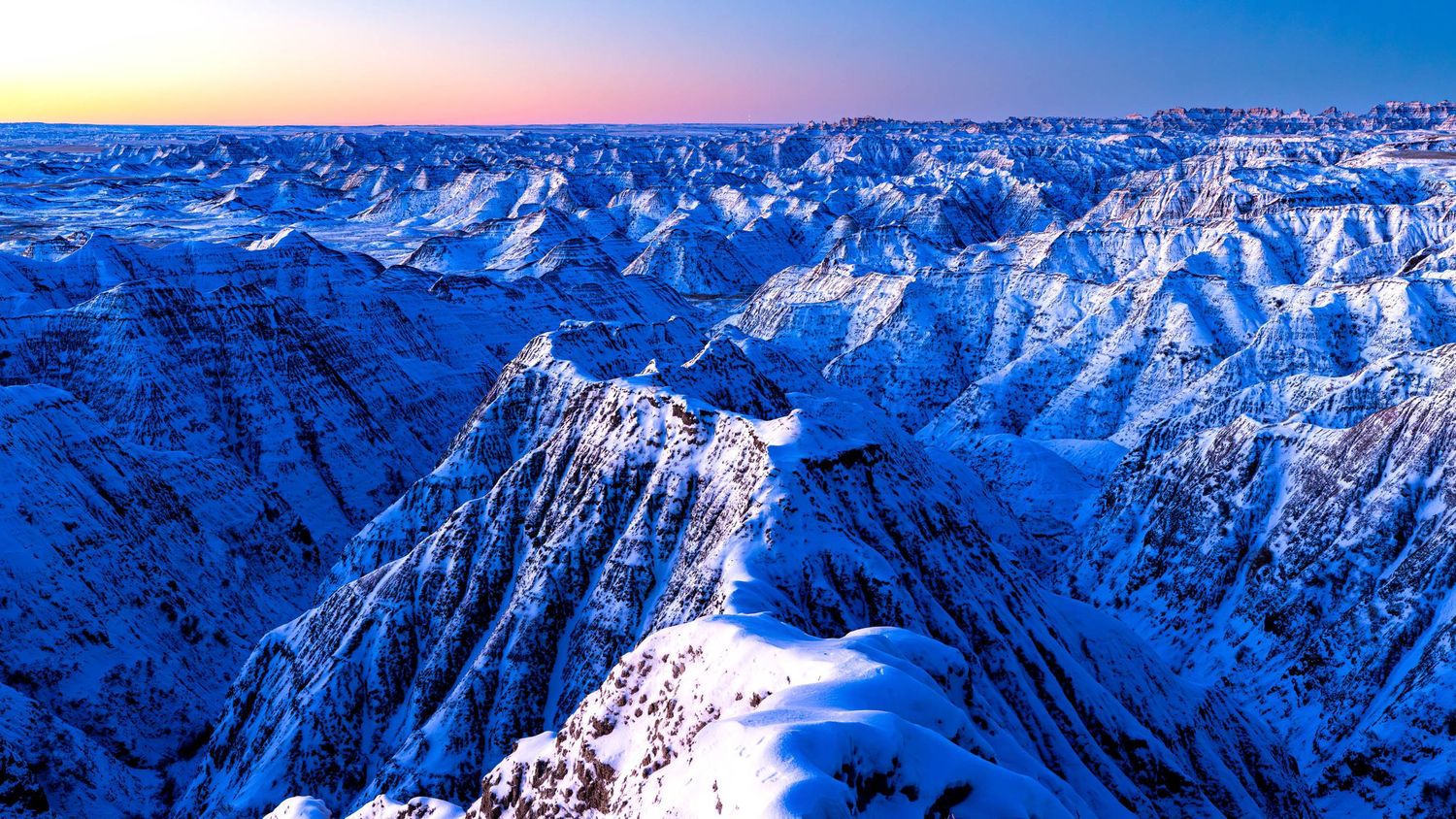 Snow-capped mountain ranges stretch across the horizon under a brilliant blue winter sky. Badlands National Park