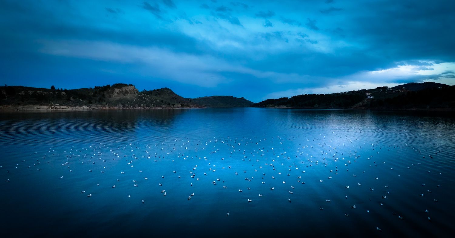 Serene blue lake panorama with mountains and dramatic clouds reflecting on calm waters.