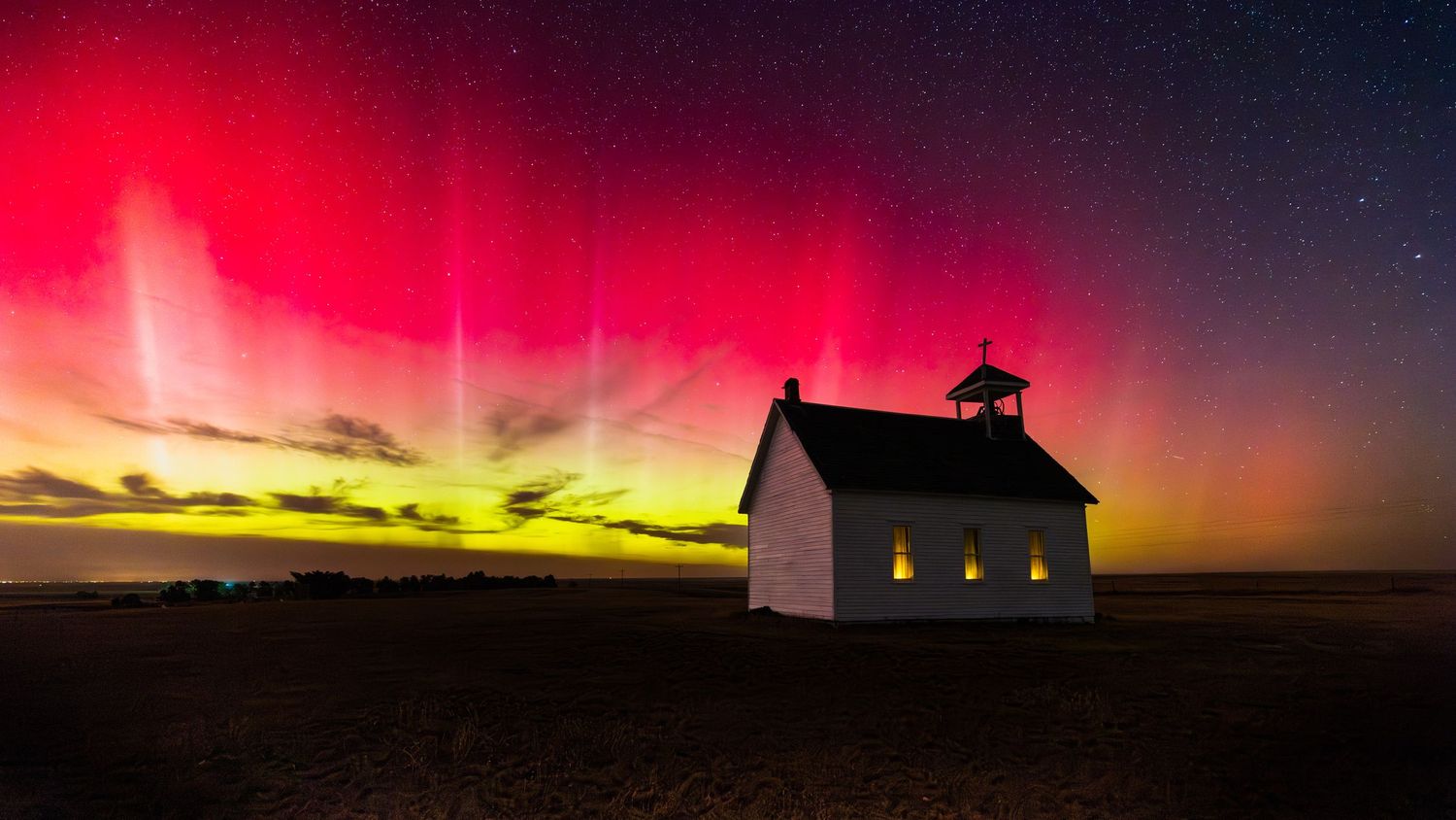 Small white church silhouetted against vibrant northern lights in night sky.