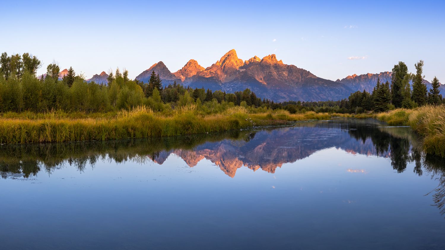 Majestic Teton mountain range reflects perfectly in calm waters of Schwabacher Landing during sunrise in Grand Teton National Park.