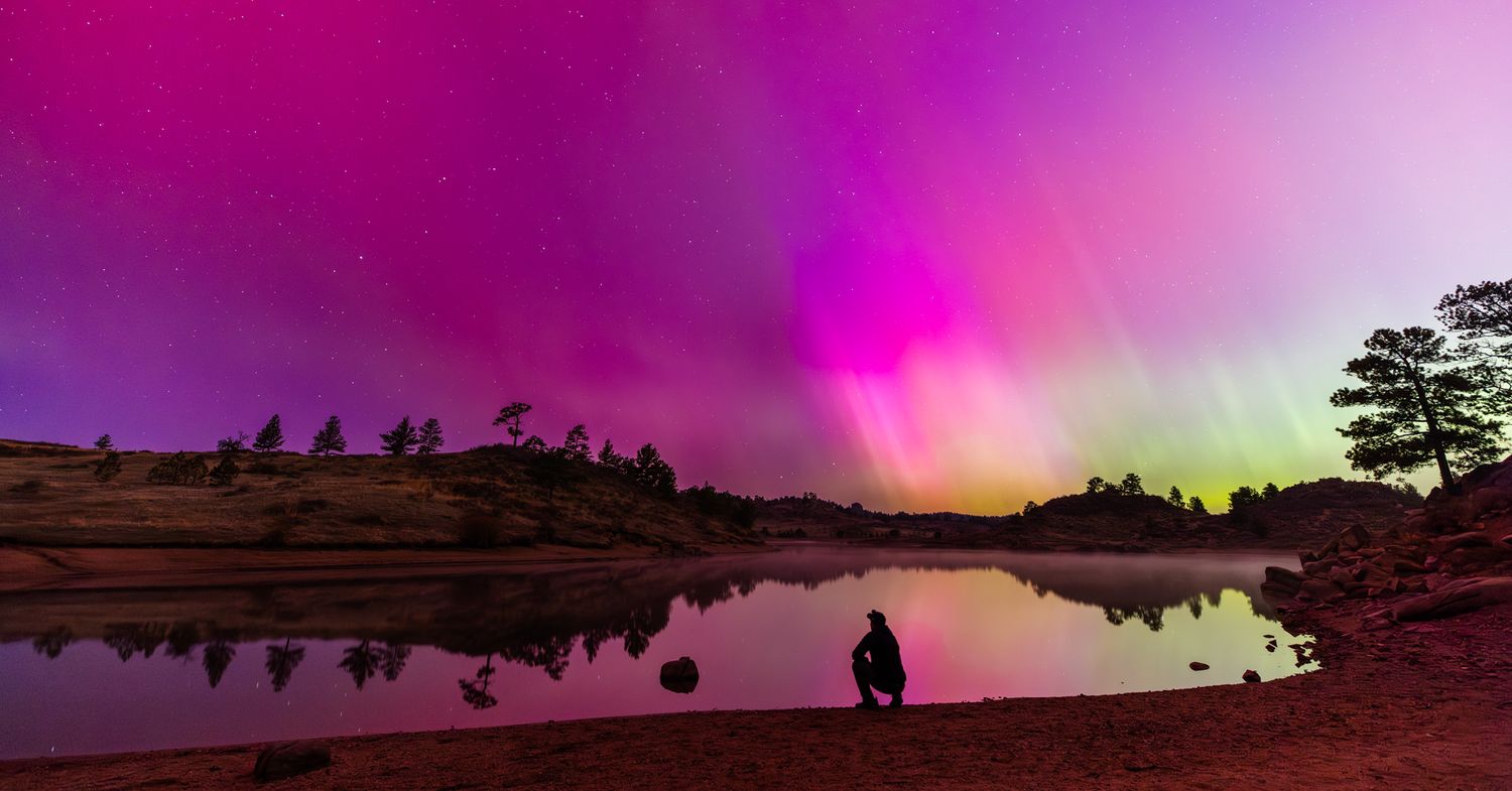 Vibrant magenta aurora sky over calm water with silhouetted figure on shoreline.