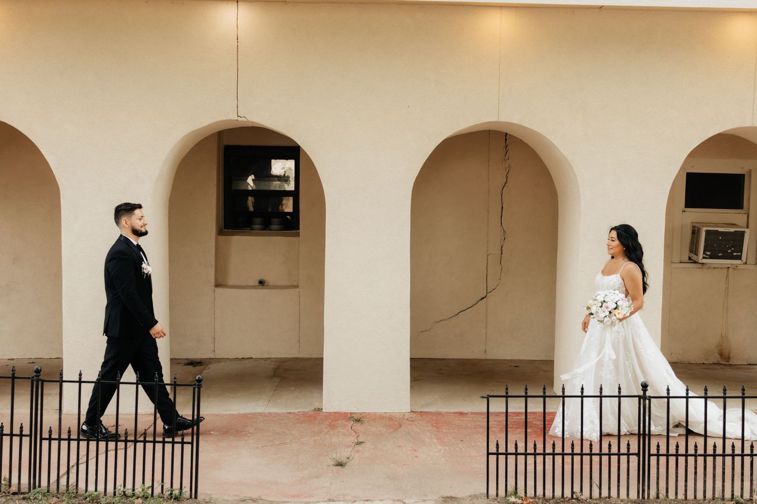 A bride and groom walk through an elegant arched corridor with soft lighting and architectural details.