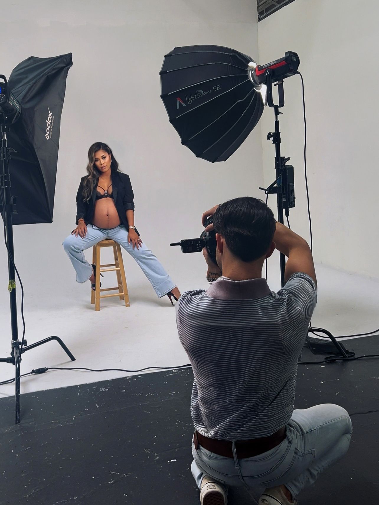 Behind-the-scenes photo shoot setup in studio showing photographer with lighting equipment and model on wooden stool.