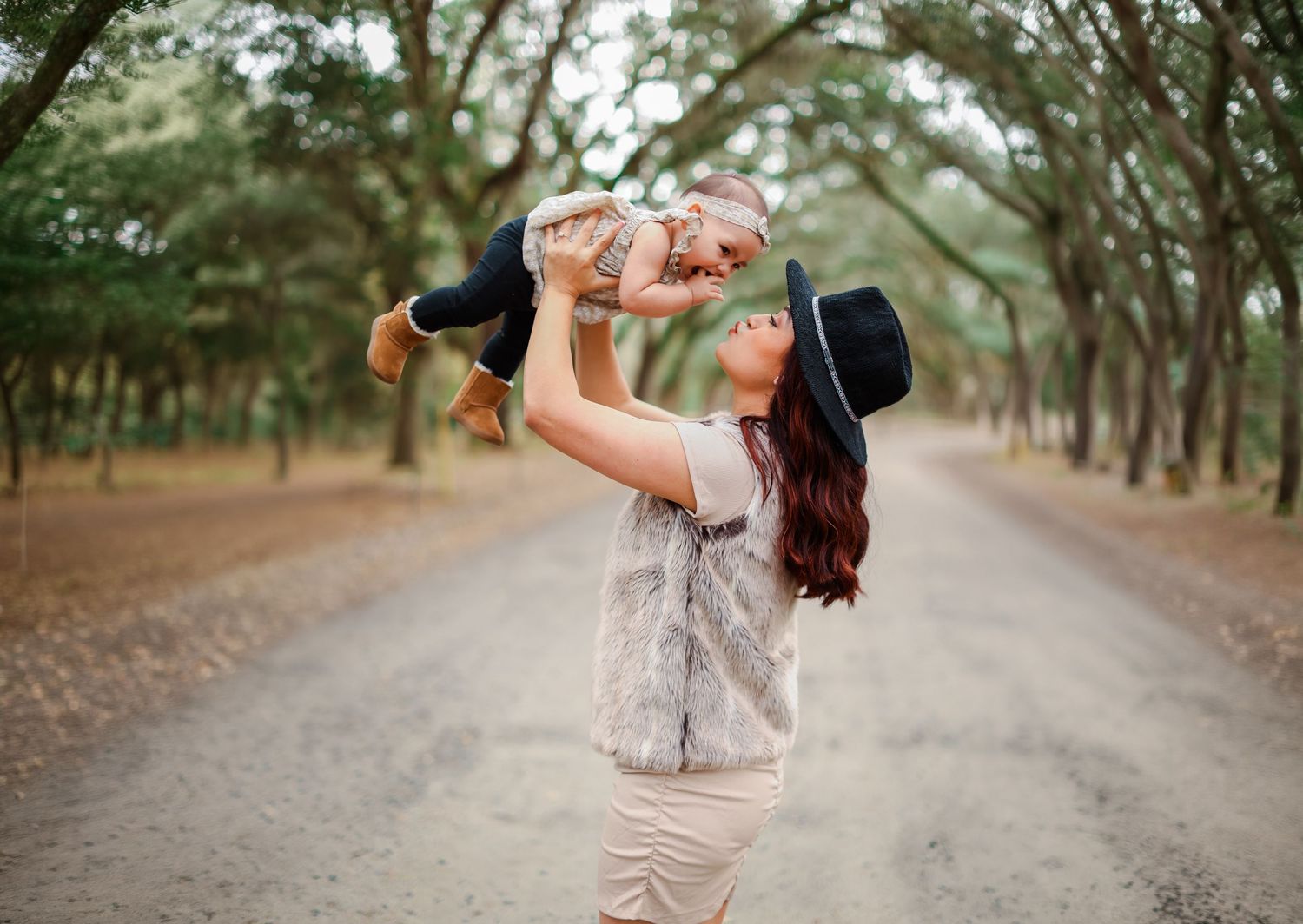 A mother joyfully lifts her baby in the air on a tree-lined dirt road, wearing a black hat.